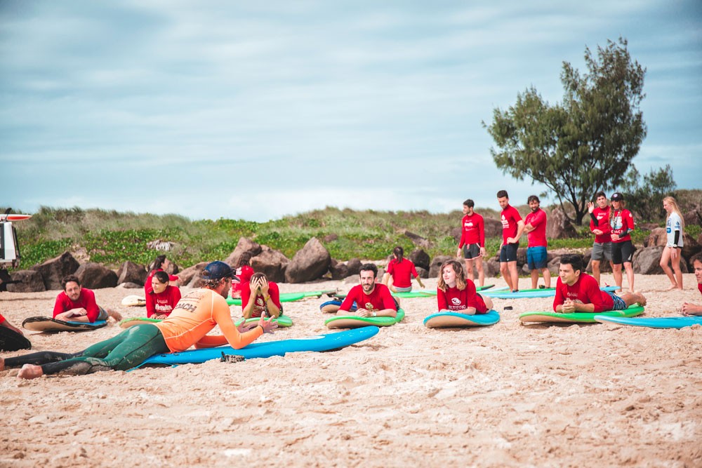 estudiantes vestidos con el uniforme de surf sentados en la arena encima de la tabla de surf. De fondo se ve la naturaleza de Australia, Gold Coast